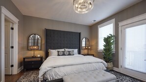 A bedroom with a large bed, two bedside tables with lamps, a chandelier, a potted plant, and a window with blinds. - Walk2Germantown Rooftop Firepit Downtown Skyline King Suite 2-Car Garage (Nashville)