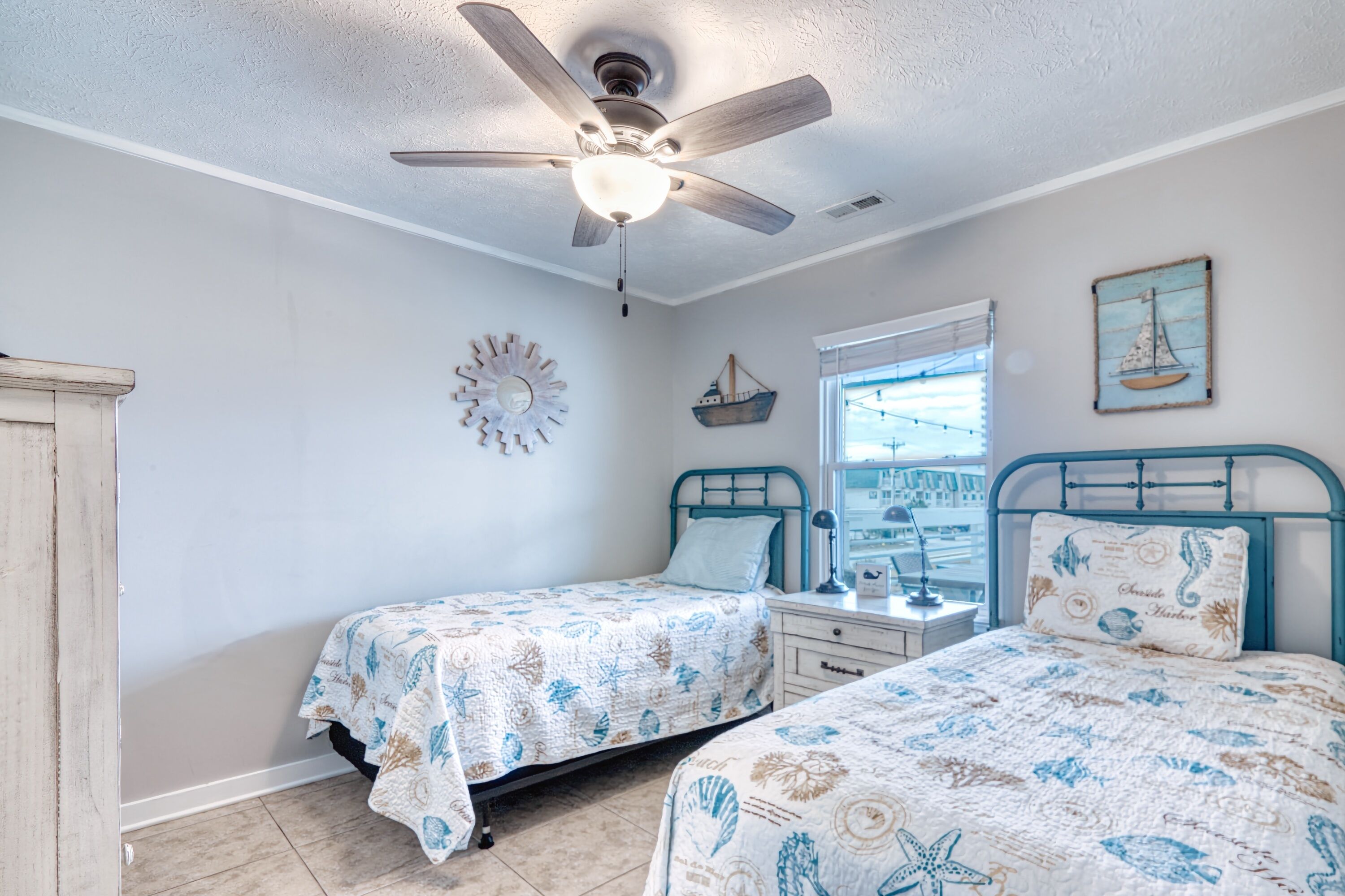 A bedroom with two beds, a ceiling fan, a window with a view, and nautical-themed decor.