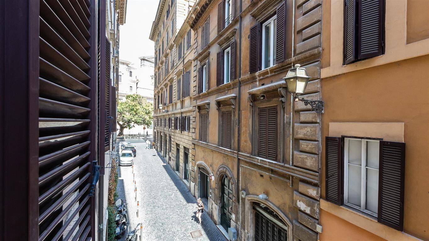 A narrow street with buildings on either side, featuring arched windows and shutters.