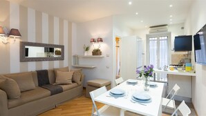 A modern living room with a sofa, a dining table set for four, and a kitchen area with a TV and appliances.