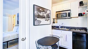 A compact kitchen with white cabinets, a microwave, a stove, and a black refrigerator. There is a small dining area with a metal chair and a table. A framed picture hangs on the wall above the kitchen counter. - 313 Room with a view 1 block from the beach (Fort Lauderdale)