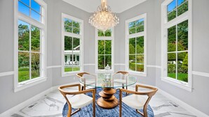 Un coin repas avec une table en verre et des chaises en bois, de grandes fenêtres donnant sur un jardin et un lustre.
