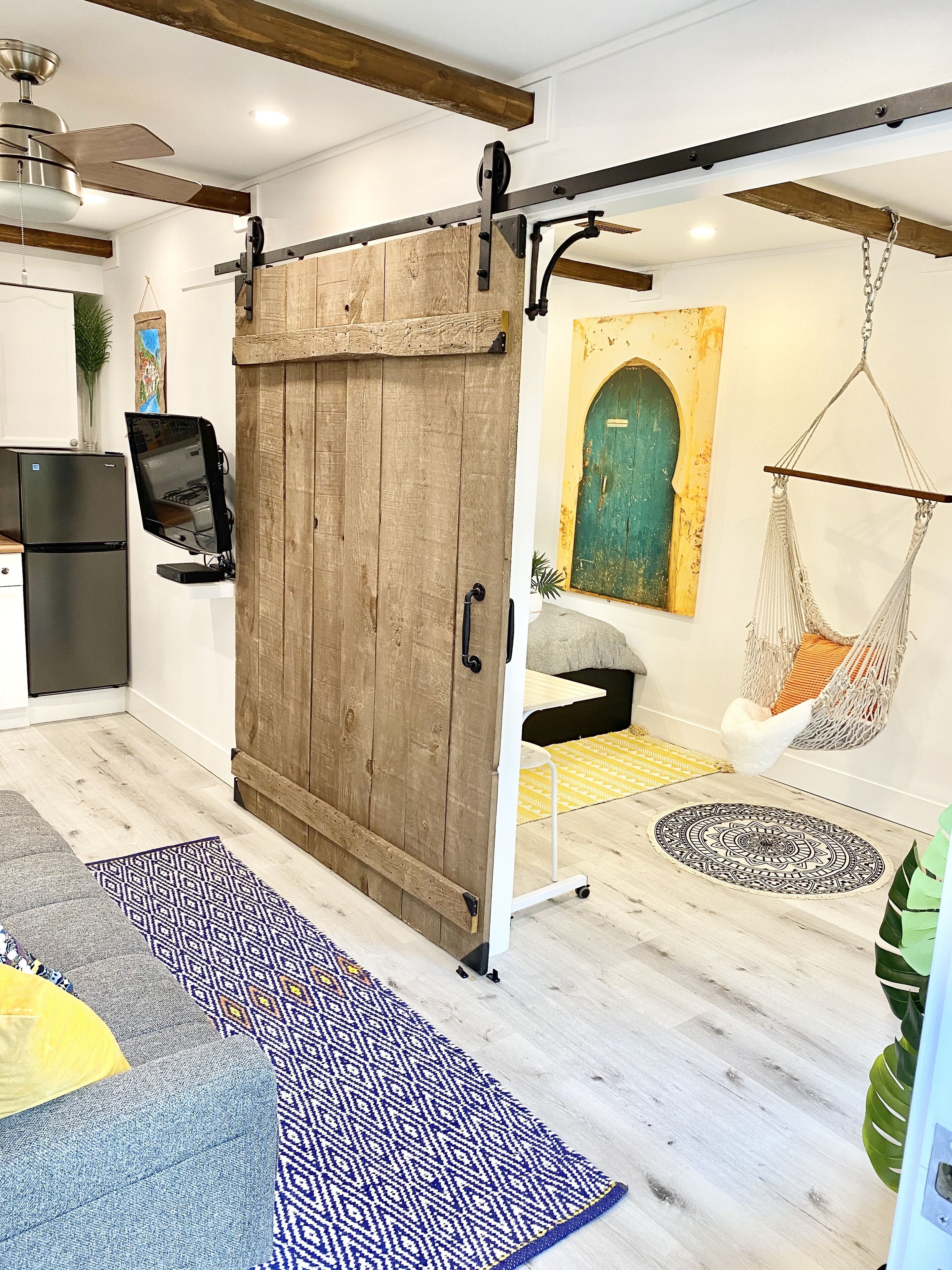 A modern living room with a sliding barn door, a hanging hammock, a flat-screen TV, and a kitchen area.