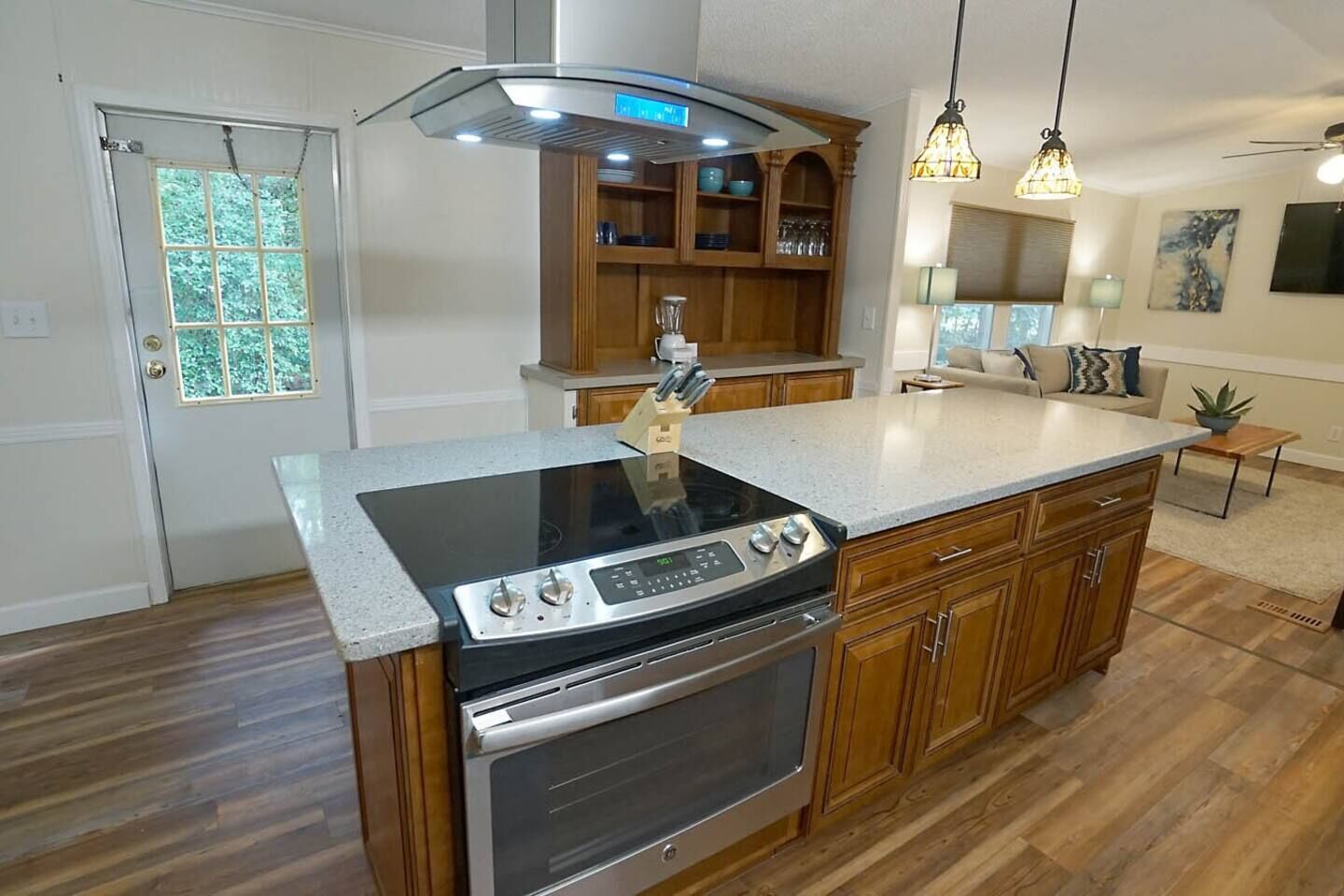 A kitchen with a central island, built-in oven, stainless steel range hood, wooden cabinets, and a window with a view of greenery.
