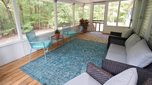 Een zonnedek met een houten vloer, een blauw gestreept vloerkleed, blauwgroene fauteuils, een grijze bank en een potplant.