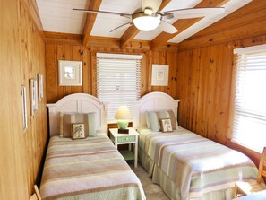 A room with two single beds, wooden walls, a ceiling fan, and a window with blinds.