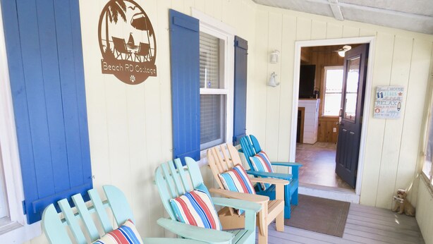 A porch with several Adirondack chairs, a sign reading "Beach RD Cottage," and a door leading to an interior space.