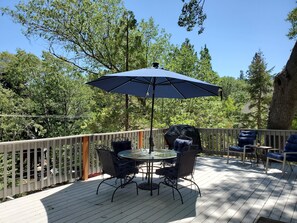 Eine Holzterrasse mit rundem Tisch, Stühlen, einem großen Sonnenschirm und einem Grill.