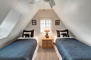 Ein kleines, gemütliches Schlafzimmer im Dachgeschoss mit zwei Betten, einem Nachttisch, einer Lampe und einem Fenster mit Jalousien.