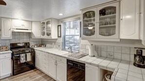 A kitchen with white cabinetry, a built-in oven, a dishwasher, and a window with blinds. - Lovely Lake View 4 BR Cabin w/ Large Kitchen & Deck. Across from lake access (Lake Arrowhead)