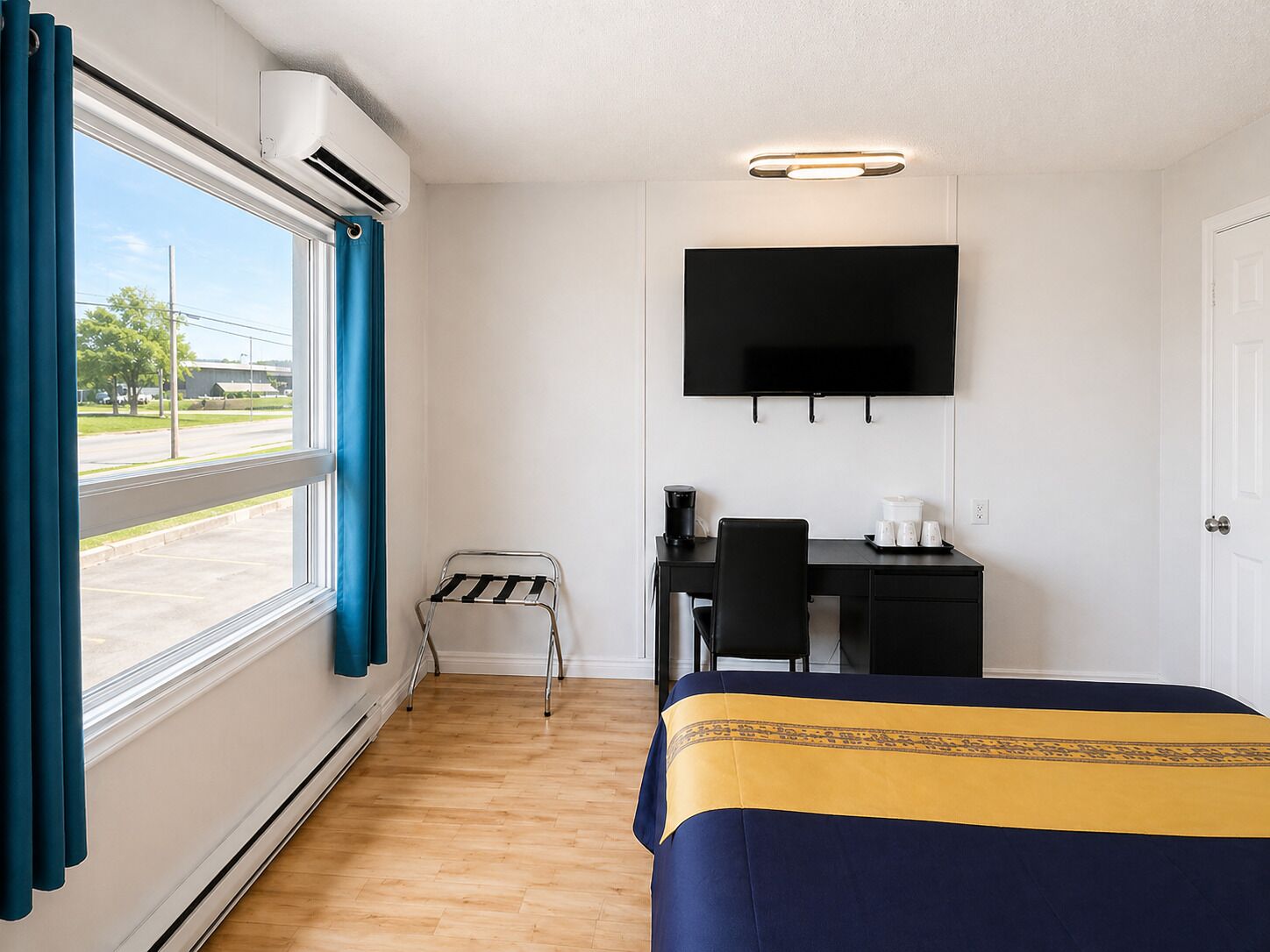 A hotel room with a bed, a desk with a chair, a TV mounted on the wall, and a window with curtains.