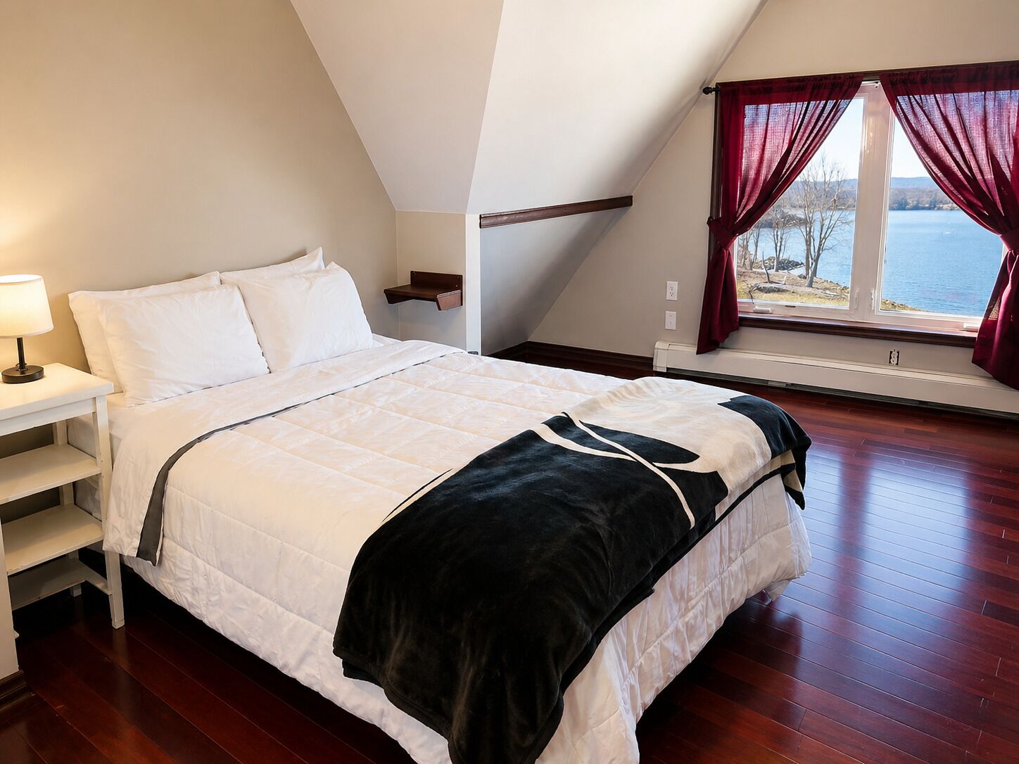 A bedroom with a bed, a nightstand, a window with curtains, and a view of the outdoors.