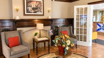 A lobby with two armchairs, a table with a lamp, a framed picture, a chandelier, and a door leading to a reception area.