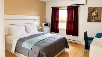 A hotel room with a bed, a desk, a chair, an air conditioner, a microwave, and a painting on the wall.
