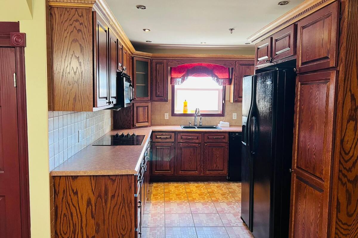 Una cocina con armarios de madera, un refrigerador negro, una estufa y un ventanal con un adorno rojo.