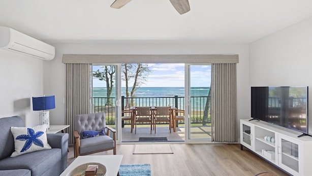 A modern living room with a sofa, a coffee table, a TV, and a balcony with a view of the sea. - RENOVATED 10/21! Corner Oceanview Condo w/ AC!! (Kapaa)