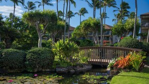 A lush garden with a wooden bridge over a pond, surrounded by palm trees and various plants. - RENOVATED 10/21! Corner Oceanview Condo w/ AC!! (Kapaa)