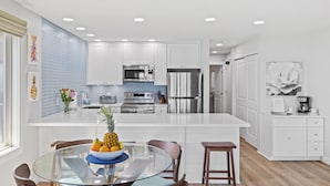A modern kitchen with white cabinets, stainless steel appliances, and a glass dining table with chairs. - RENOVATED 10/21! Corner Oceanview Condo w/ AC!! (Kapaa)