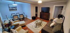 Un salon comprenant un canapé, un pouf, une table basse et un téléviseur posé sur un meuble. On y trouve également un coin repas avec une table et des chaises.