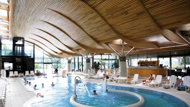 Una piscina interna con soffitto in legno, sedie a sdraio e un’area bar.