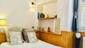 Una camera da letto con una testiera in legno, un letto con biancheria da letto bianca e cuscini decorativi, un comodino con una lampada, uno scaffale con libri e un vaso, e una finestra con tende.