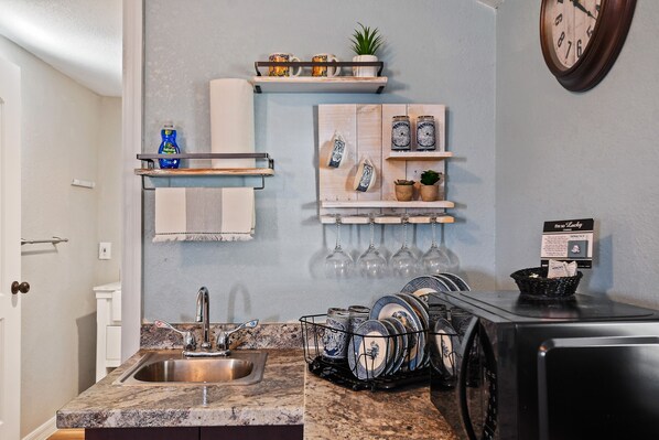 A kitchen with a granite countertop, stainless steel sink, and a microwave. There are shelves with decorative items and a clock on the wall. - Adorable guest suite with private pool and entrance. (Lake Alfred)