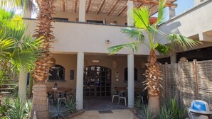 A covered outdoor seating area with tables and chairs, surrounded by palm trees and a thatched roof structure.