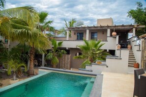 Una propiedad residencial con piscina, palmeras y un edificio de dos pisos con techo plano.