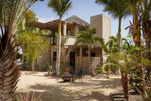 A two-story building with a thatched roof, surrounded by palm trees and a sandy courtyard.