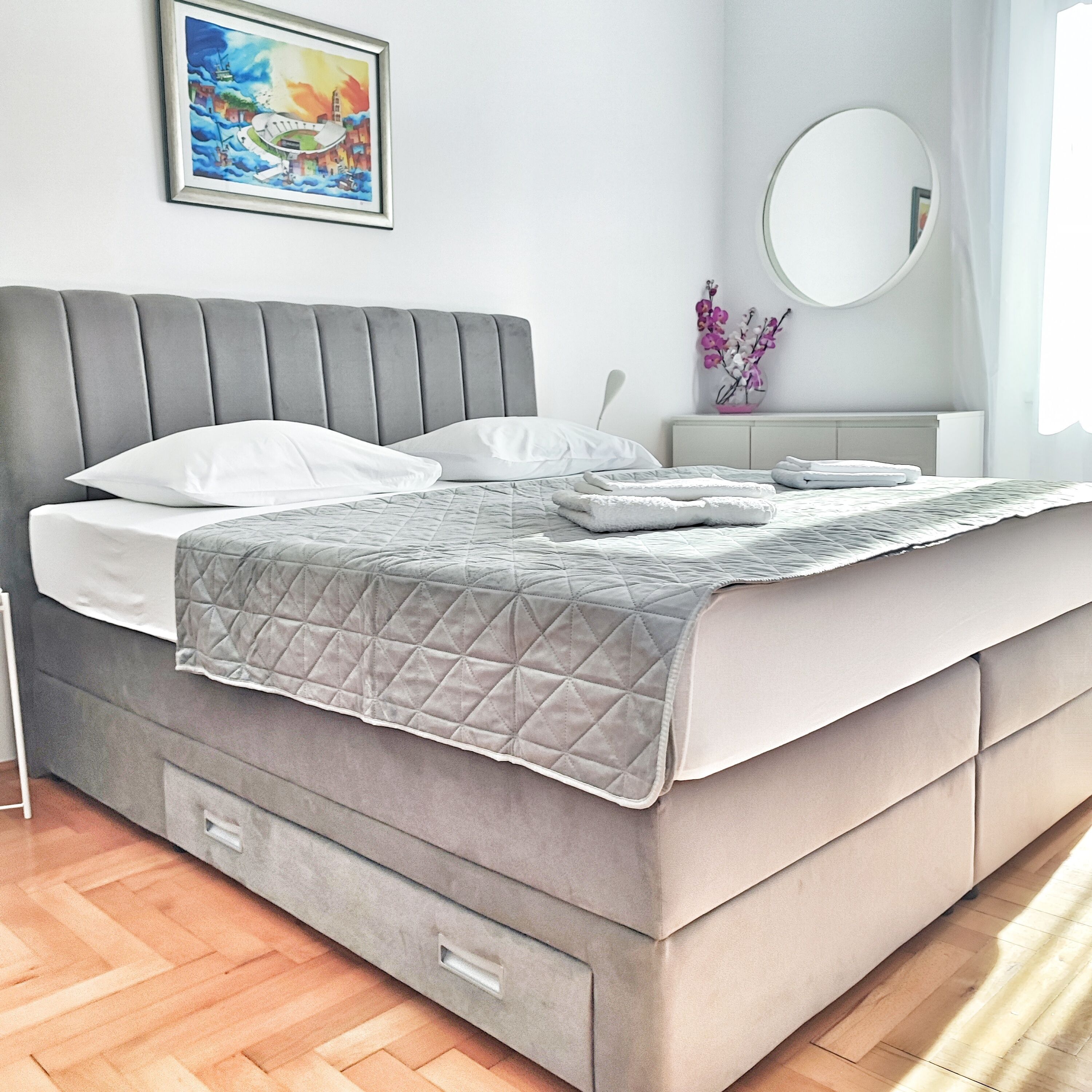 A modern bedroom with a grey upholstered bed, a quilted grey blanket, a bedside table with a vase of flowers, and a painting on the wall.