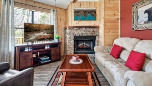 Un salon chaleureux avec une cheminée en pierre, des murs en bois, un téléviseur et un canapé agrémenté de coussins rouges.