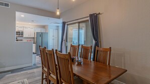 Comedor con mesa y sillas de madera, cocina al fondo y vista a través de una puerta de cristal.