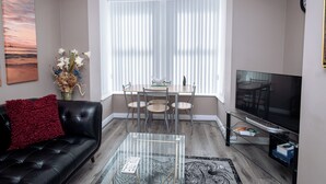 A living room with a black leather sofa, a glass coffee table, a dining table with chairs, a TV, and a large window with blinds. - Modern 1-Bed Apartment – 3 Min Walk to HOST & Pitman, Leeds (Leeds)