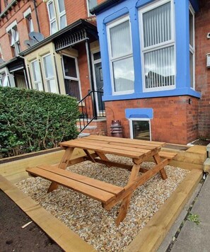 A wooden picnic table set on gravel in front of a blue-painted townhouse with a brick facade and satellite dishes. - Modern 1-Bed Apartment – 3 Min Walk to HOST & Pitman, Leeds (Leeds)