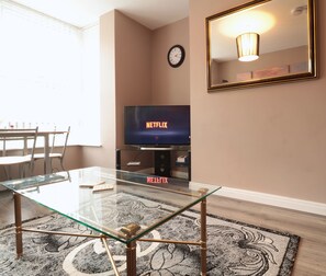 En stue med et glasbord, et fladskærms-tv der viser Netflix, et ur og et spejl på væggen.