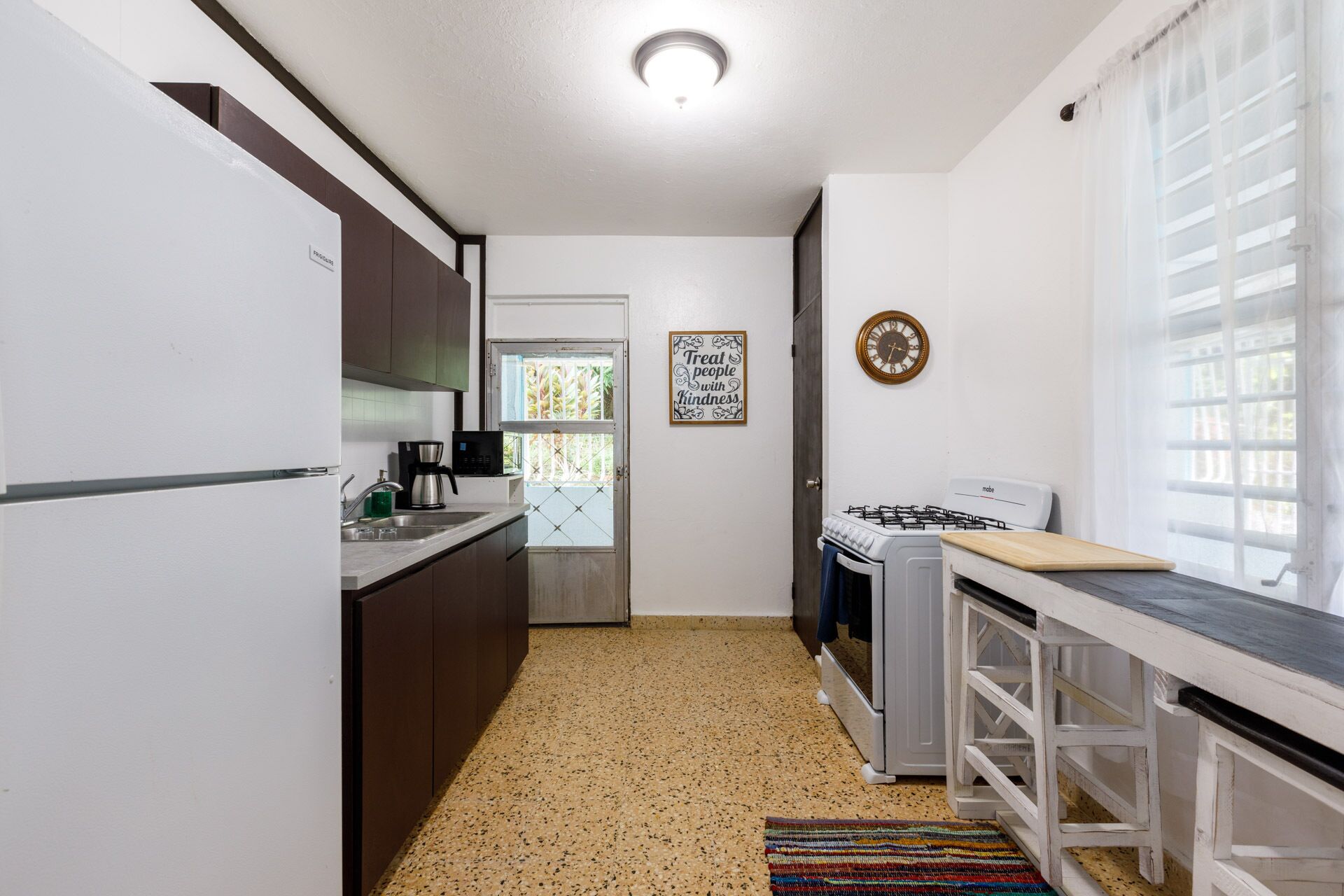 A kitchen with a stove, sink, and cabinets. There is a window with curtains and a door leading to another room.