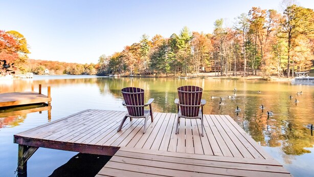 Un muelle de madera con dos sillas Adirondack mirando a un lago tranquilo rodeado de árboles de colores otoñales.