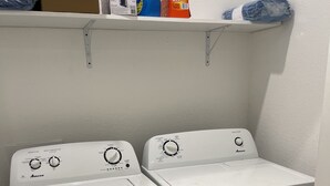 Un espace buanderie équipé de deux machines à laver à chargement frontal et d’une étagère au-dessus, où sont rangés les produits de nettoyage.