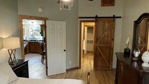 A bedroom with a bed, a dresser, and a desk. There is a door leading to a kitchen and a bathroom.