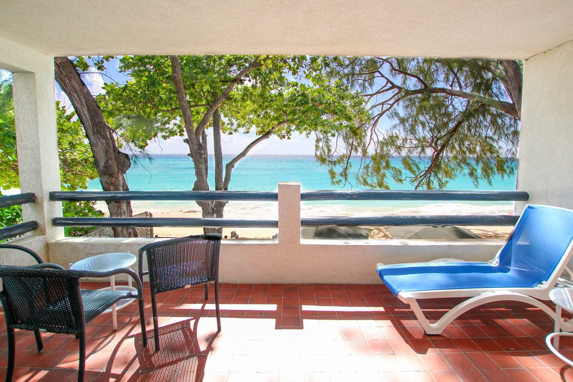 A balcony with a view of the beach, a blue lounge chair, and two wicker chairs.