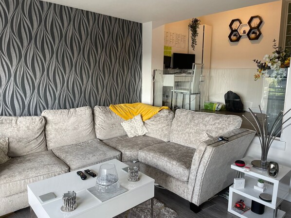 A modern living room with a grey patterned wallpaper, a grey sofa with a yellow throw, a white coffee table with decorative items, and a white side table with a vase and a plant.