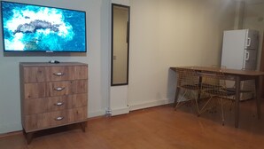 Ein Zimmer mit einem hölzernen Schminktisch, einem an der Wand befestigten Fernseher, einem Spiegel, einem Esstisch mit Drahtstühlen und einem Kühlschrank.