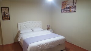 Ein ordentlich bezogenes Bett mit weißen Leinen und einem Kopfteil mit Diamantmuster. Ein kleiner Nachttisch steht daneben, mit einer Lampe. An der linken Wand hängt ein gerahmtes Bild.