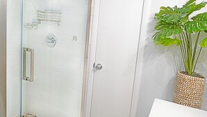 A bathroom with a glass shower enclosure, a white door with a silver handle, and a potted plant with broad leaves. - Cheerful 2 Bedroom Townhome With Fireplace & Private Patio  in New Smyrna Beach! (New Smyrna Beach)