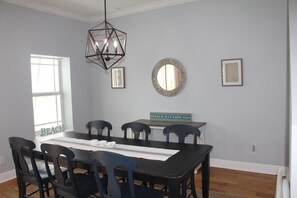 Uma sala de jantar com uma mesa de madeira escura, seis cadeiras, um lustre pendente, um espelho redondo e uma placa emoldurada na parede.