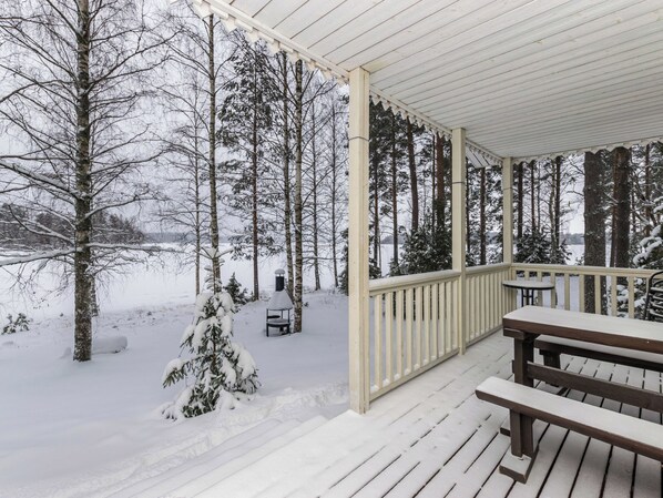 A covered outdoor deck with a wooden bench and a table, surrounded by snow-covered trees.
