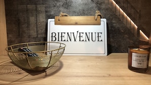 A welcome sign, a candle, and a basket with remote controls on a wooden surface.