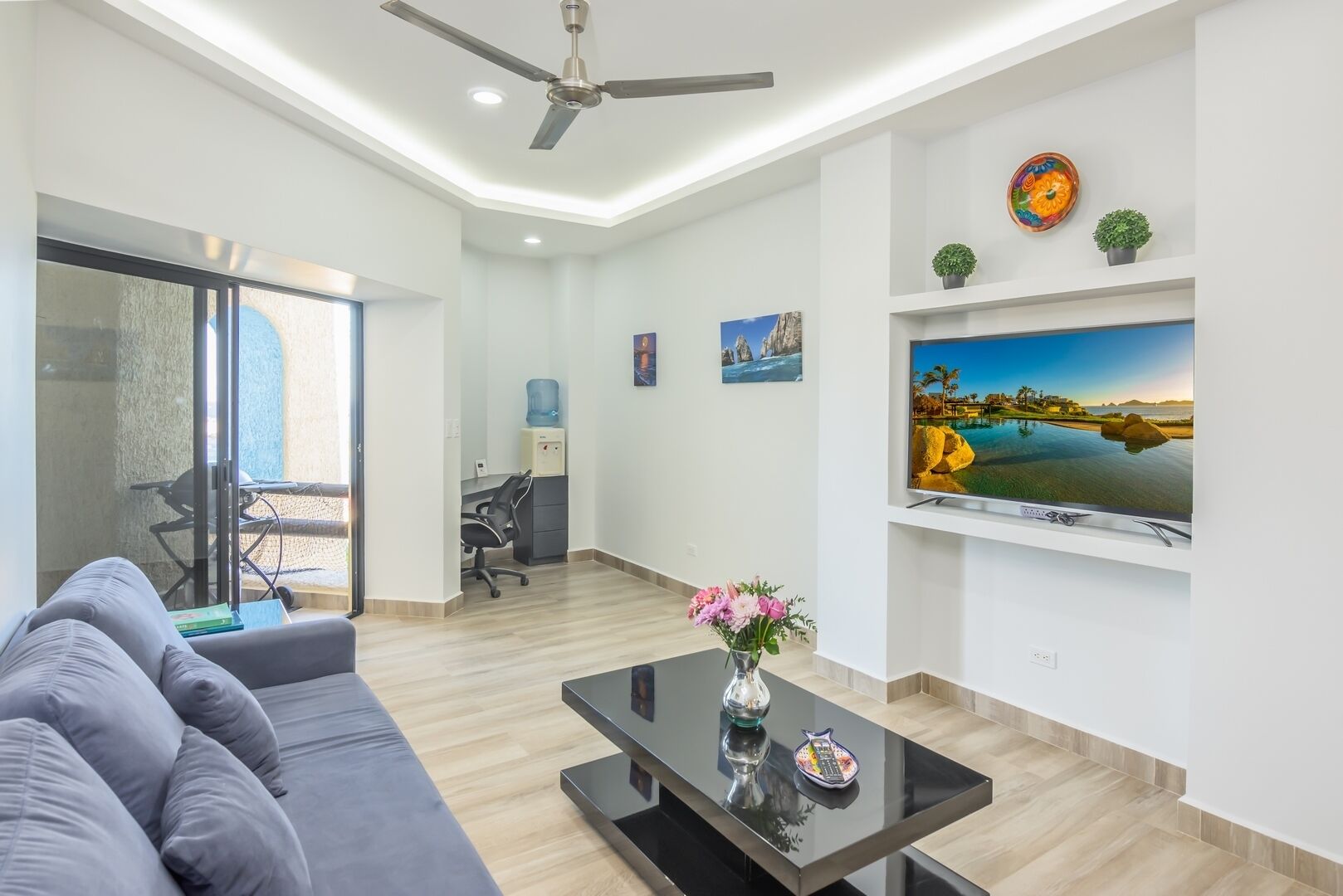 A modern living room with a sofa, a coffee table with a vase of flowers, a TV on the wall, and a ceiling fan.