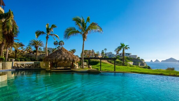 A clear blue pool with a thatched-roof structure, palm trees, and a view of the sea. - AWESOME OCEAN VIEWS, GREAT LOCATION, PERFECT VACATION (Cabo San Lucas)