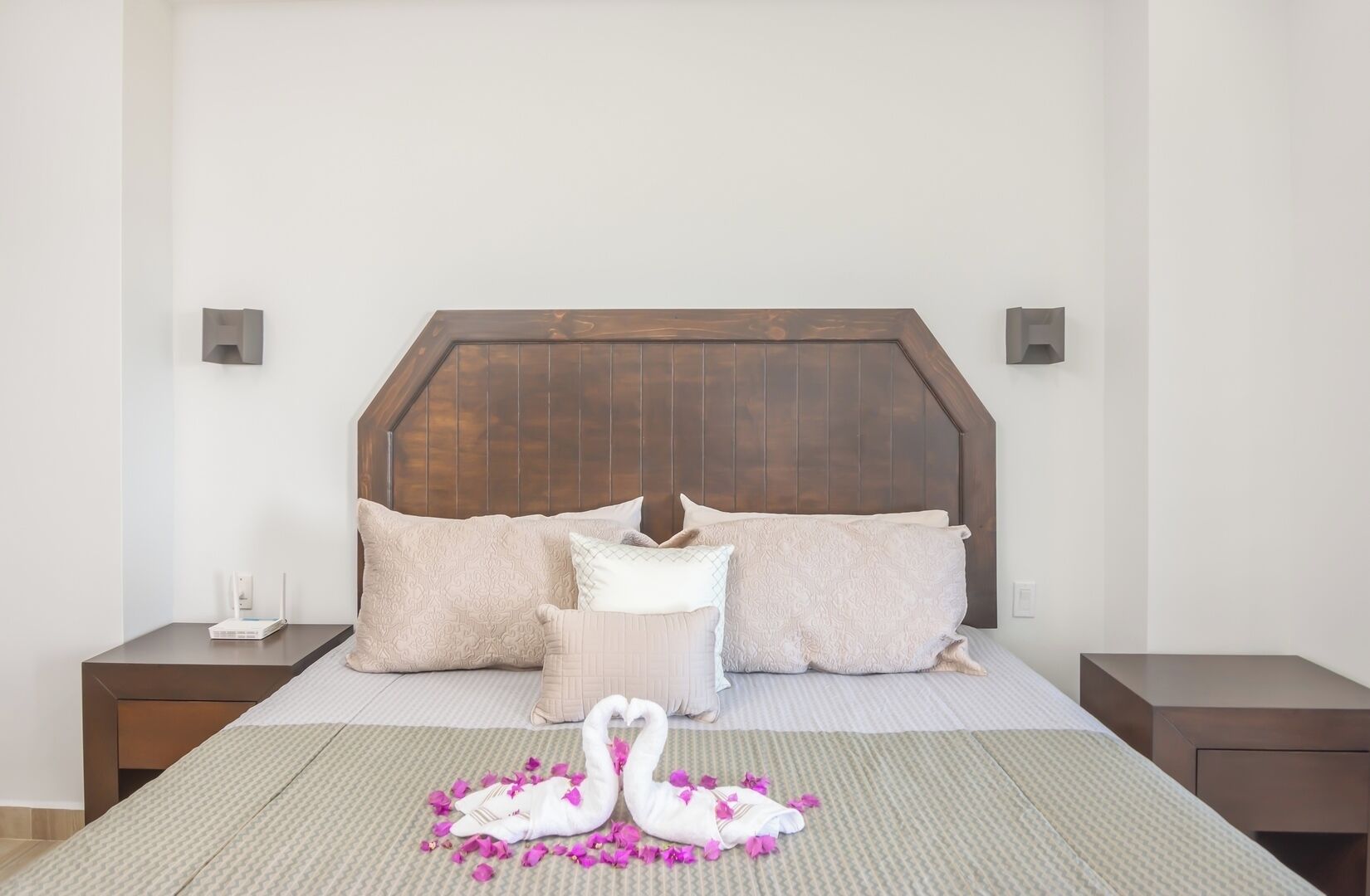 Een slaapkamer met een houten hoofdbord, een bed met meerdere kussens, een nachtkastje met een lamp, en een decoratieve zwaan en bloemen op het bed.